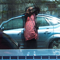 A woman in a rich peach dress and black cardigan, holding a camera 
 to her eyes taking a picture of herself in a reflective window, showing a reflections that's distorted and layered,
 a blue car parked behind her.